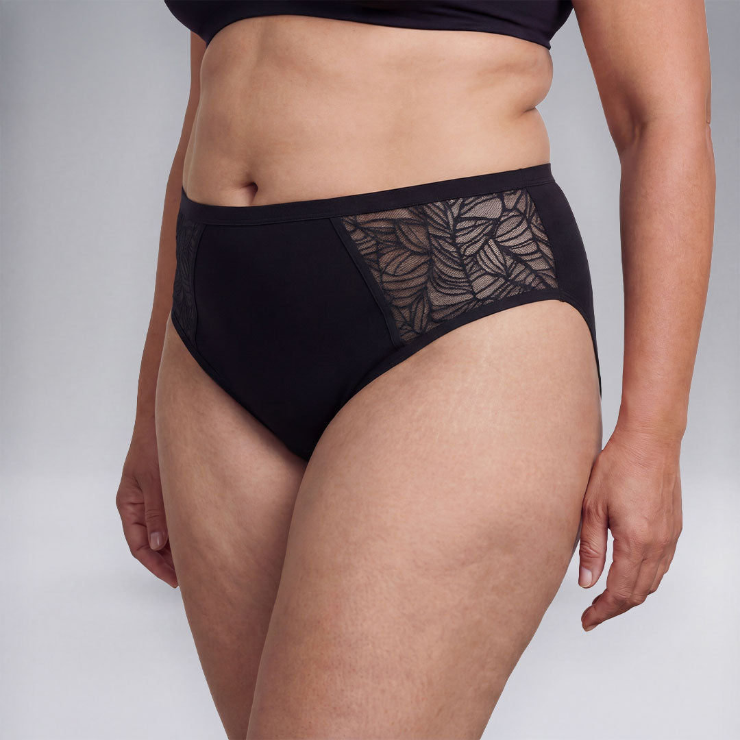 Femme portant une culotte absorbante noire avec empiècements en dentelle sur les côtés, solution lavable et discrète pour incontinence féminine, protection confortable et respirante contre les fuites urinaires légères à modérées.